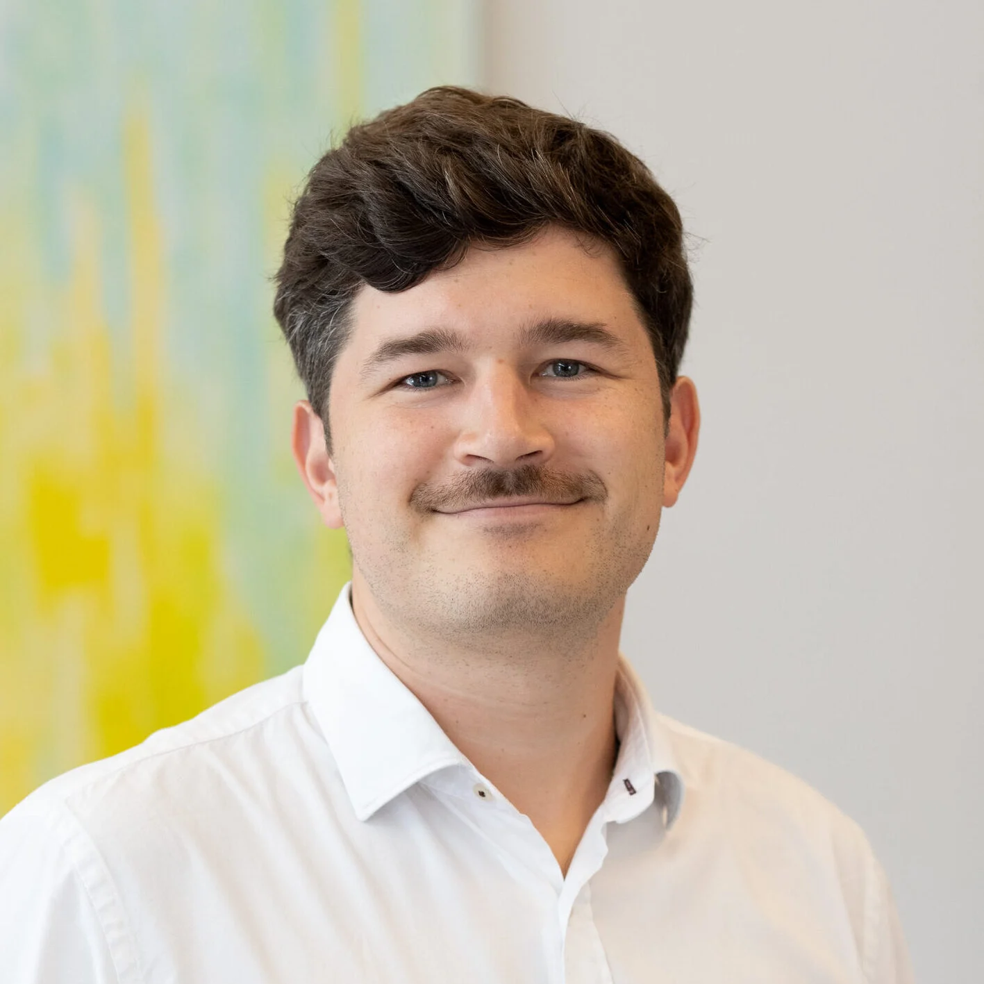 Smiling man with dark hair and a mustache in a white shirt; ‘SCS Contact’ is blurred against a colorful background. Smiling man with dark hair and a mustache in a white shirt; 'SCS Contact' is blurred against a colorful background.