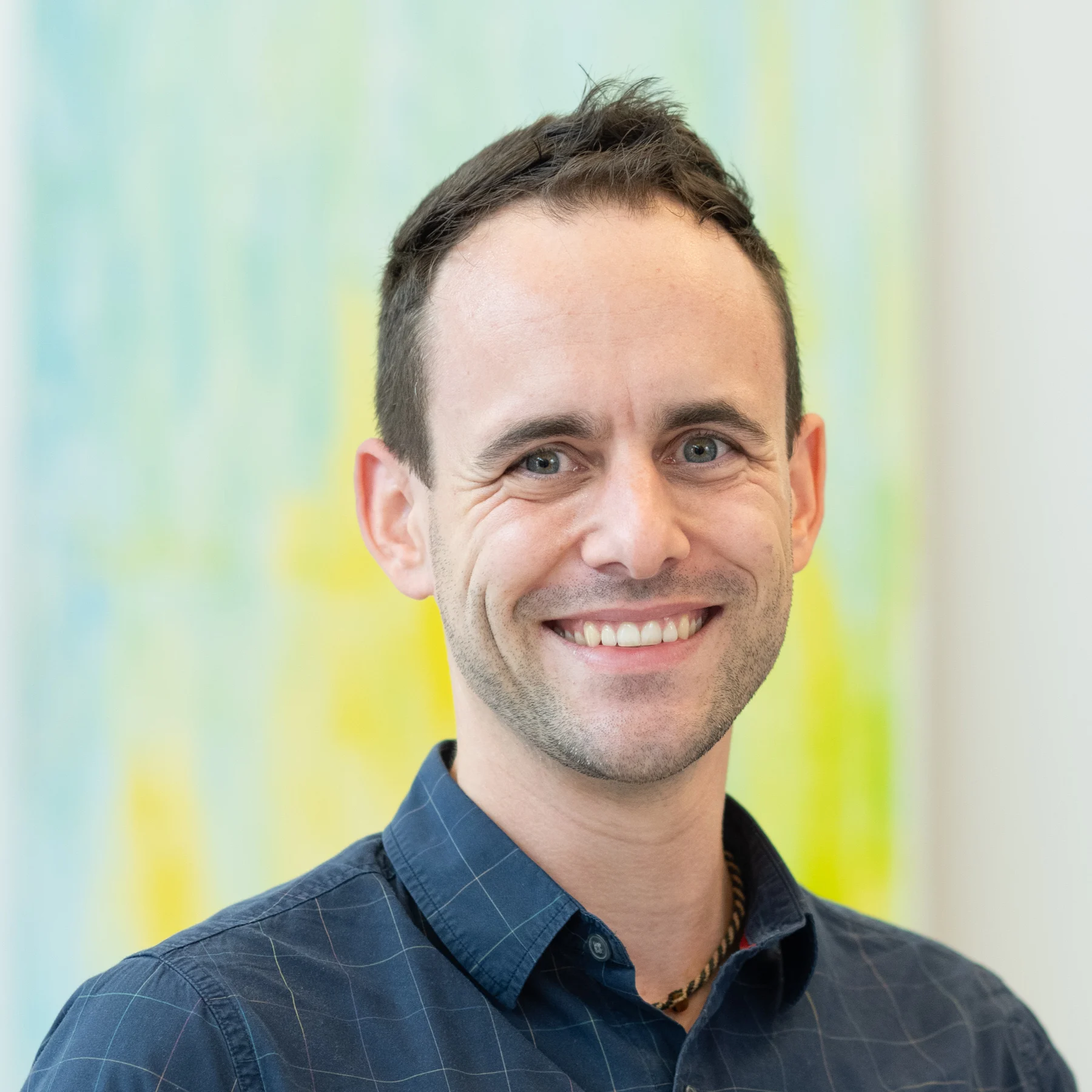 A man in a blue shirt smiles, in front of a blurred pastel blue and yellow background with the inscription “scs kontakt”. A man in a blue shirt smiles, in front of a blurred pastel blue and yellow background with the inscription "scs kontakt".