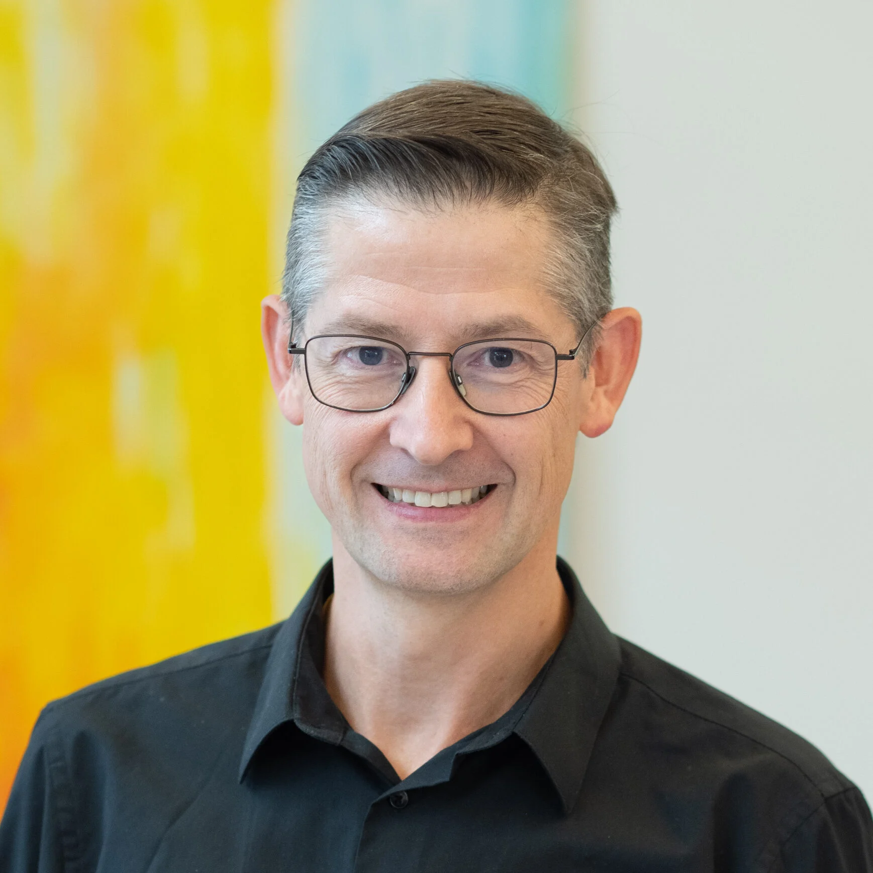 Smiling man with glasses and short hair in a black shirt, colorful background, member of the SCS team. Smiling man with glasses and short hair in a black shirt, colorful background, member of the SCS team.