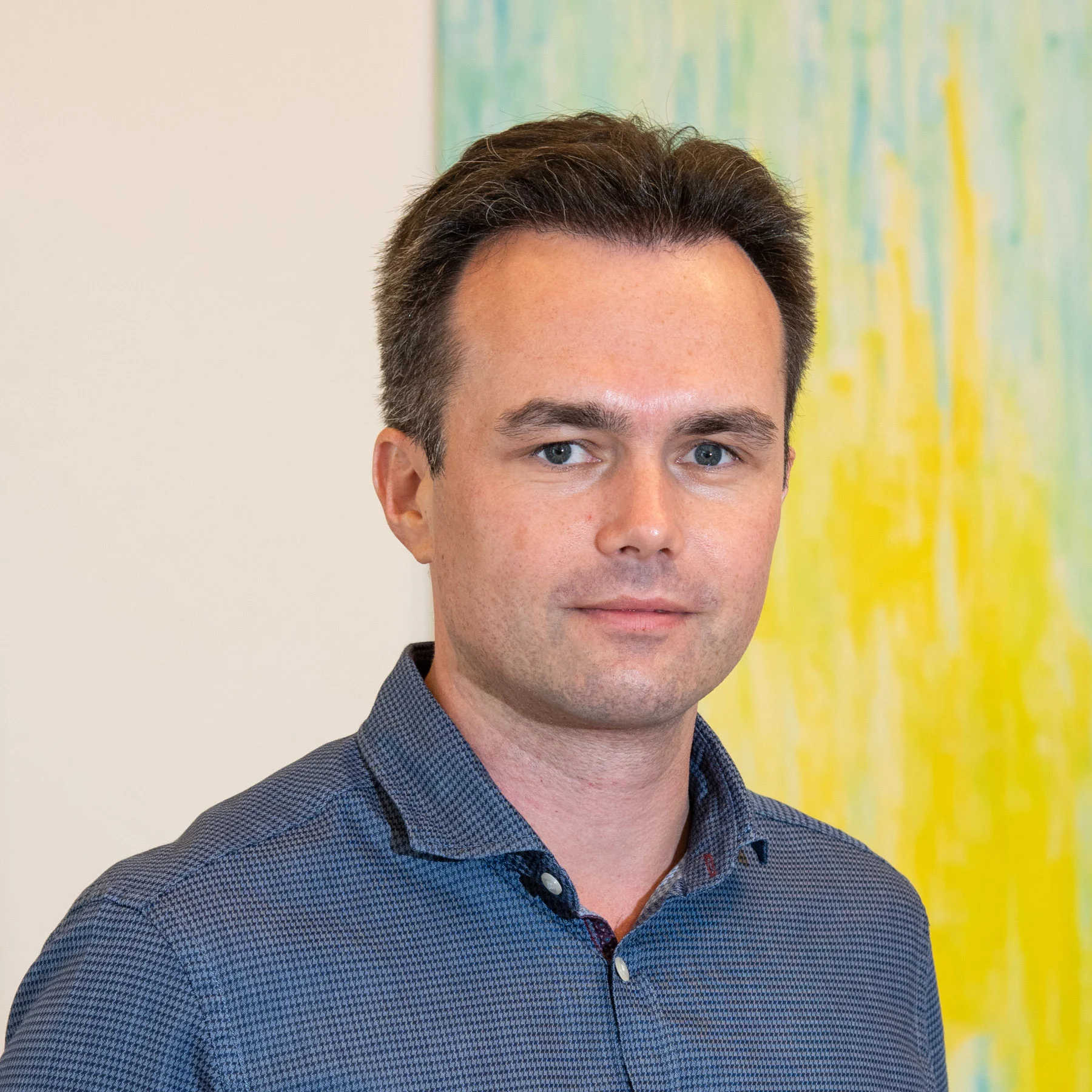 A man in a blue shirt, a member of the SCS team, stands in front of a colorful yellow-blue background. A man in a blue shirt, a member of the SCS team, stands in front of a colorful yellow-blue background.
