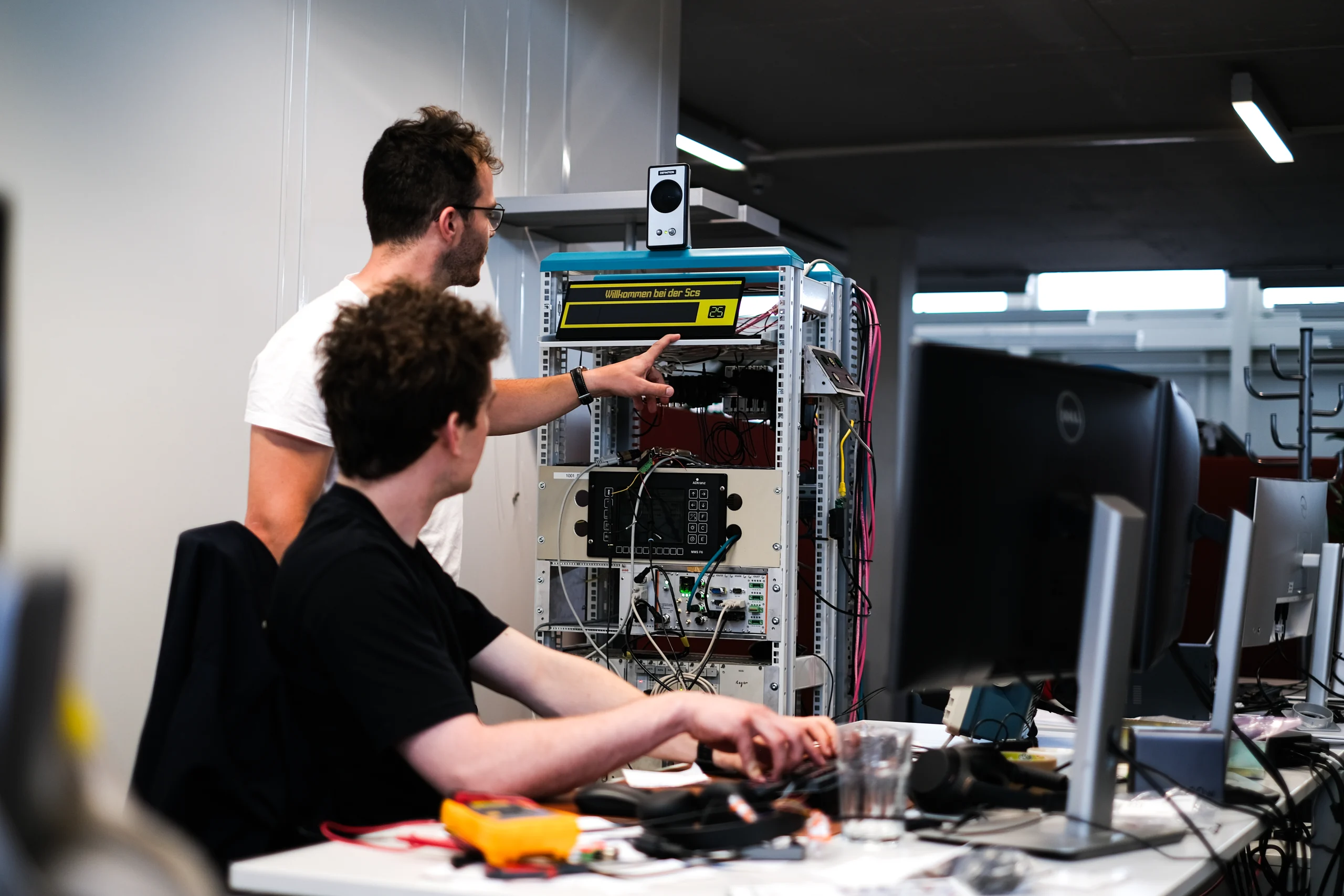 Zwei Entwickler an einem offenen Testaufbau mit verkabeltem Rack, Anzeigemodul und Messgeräten im Büro.