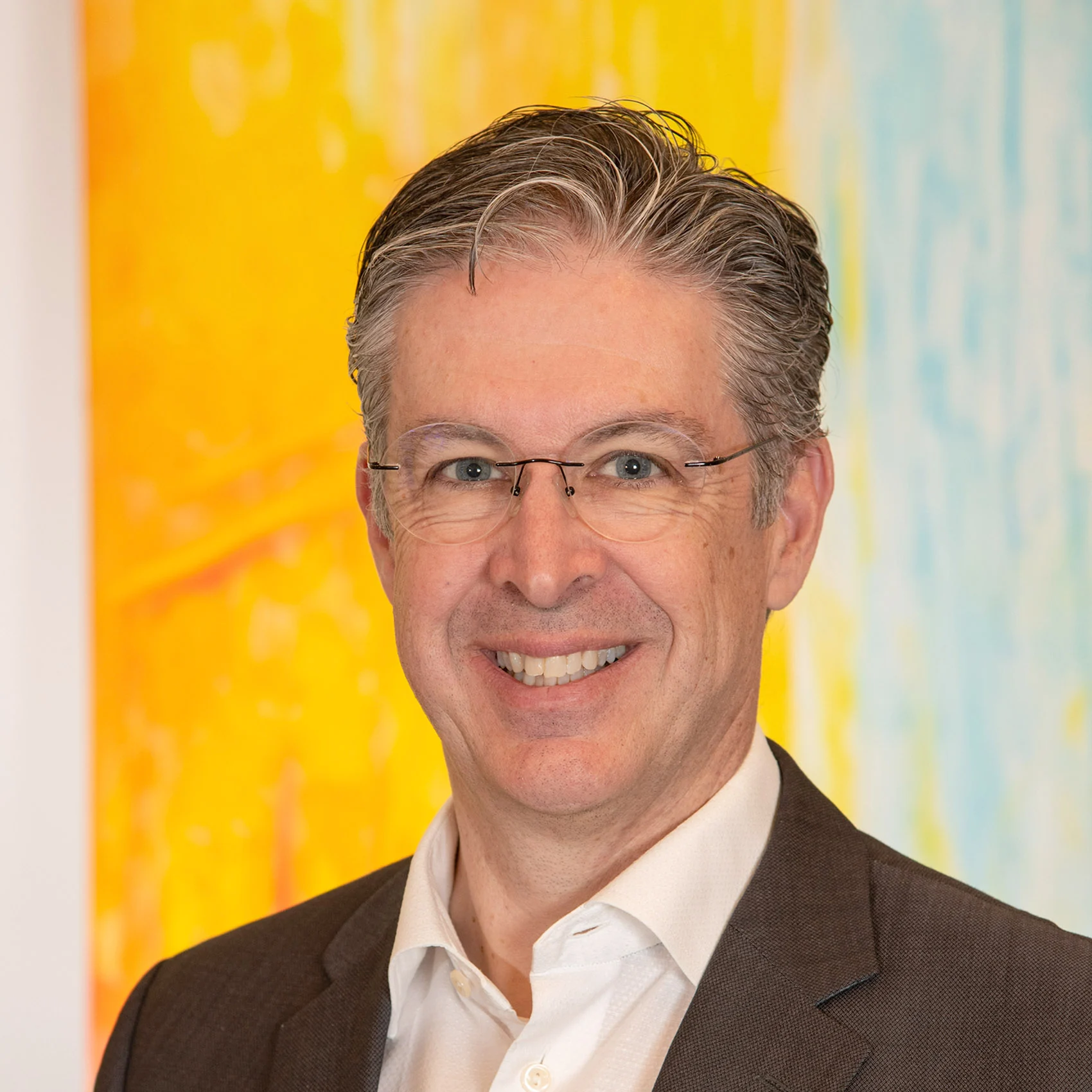 A smiling man in a suit and glasses stands against a bright background, representing the SCS team. A smiling man in a suit and glasses stands against a bright background, representing the SCS team.