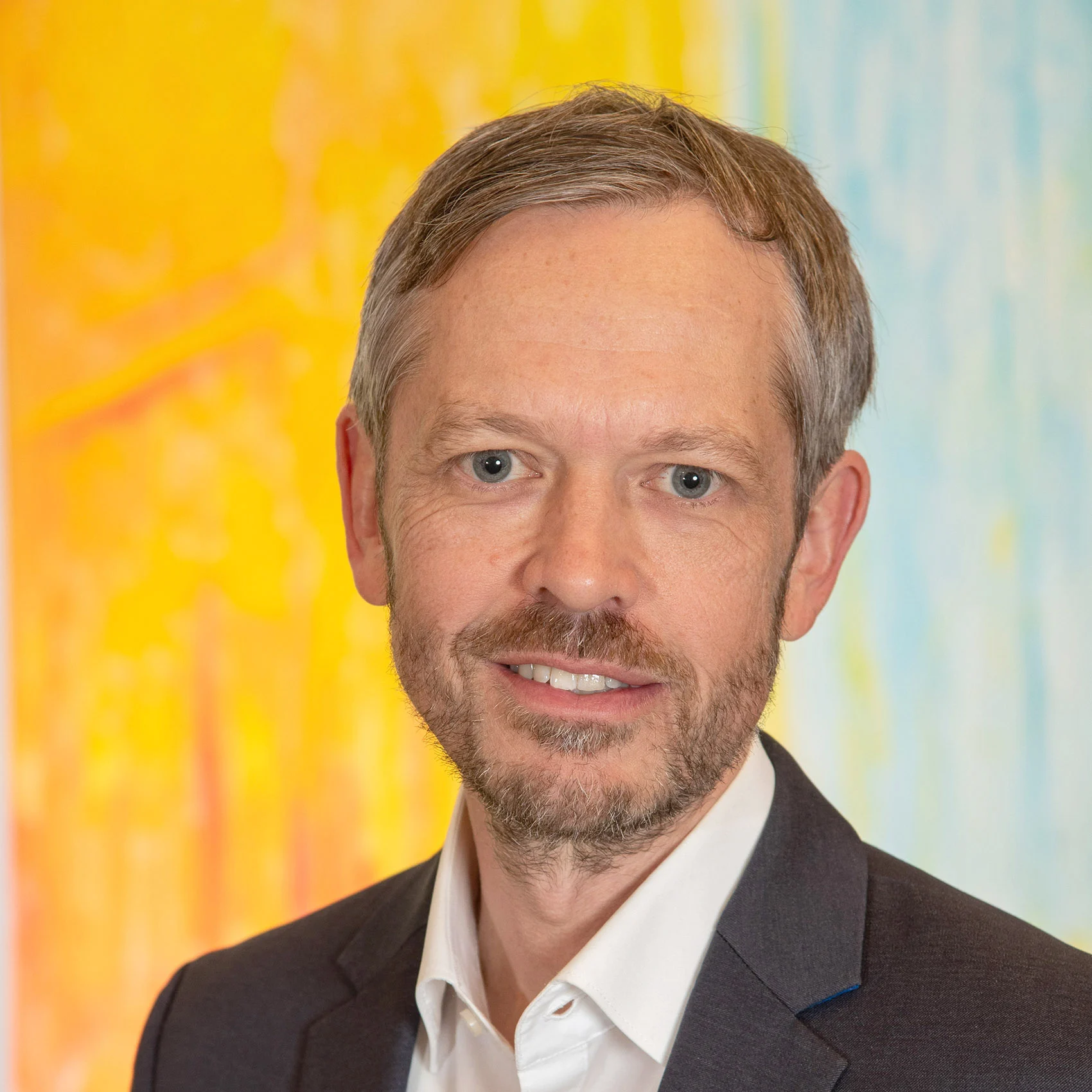 A man in a suit, a member of the SCS team, smiles in front of a colorful yellow-blue background. A man in a suit, a member of the SCS team, smiles in front of a colorful yellow-blue background.
