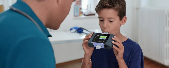 A boy blows into a medical device, strengthening his digital resilience, while an adult watches him in a bright room.