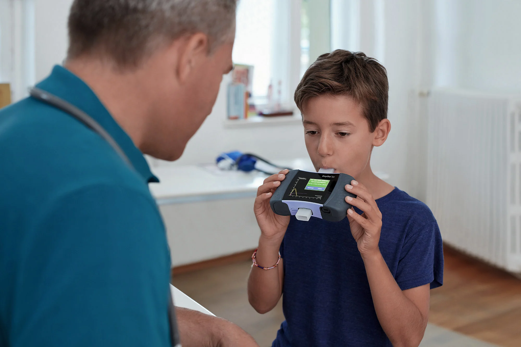 A boy blows into a medical device, strengthening his digital resilience, while an adult watches him in a bright room.