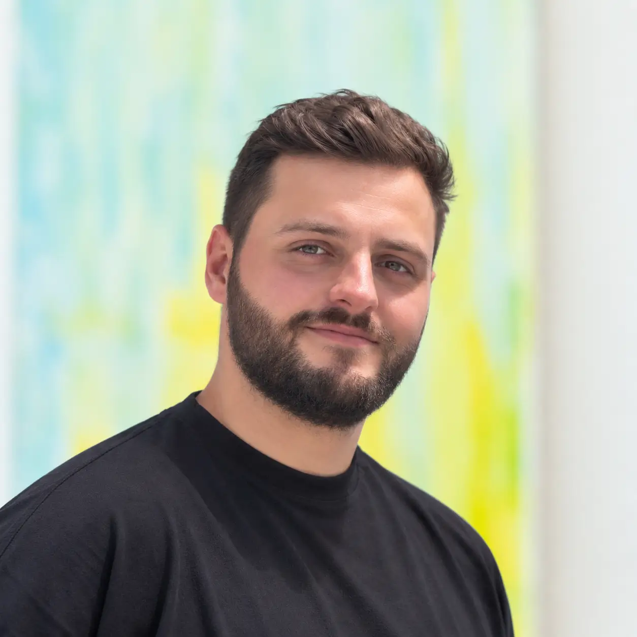 A man with a beard and black shirt stands smiling, ready for your SCS contact, with pastel tones in the background.