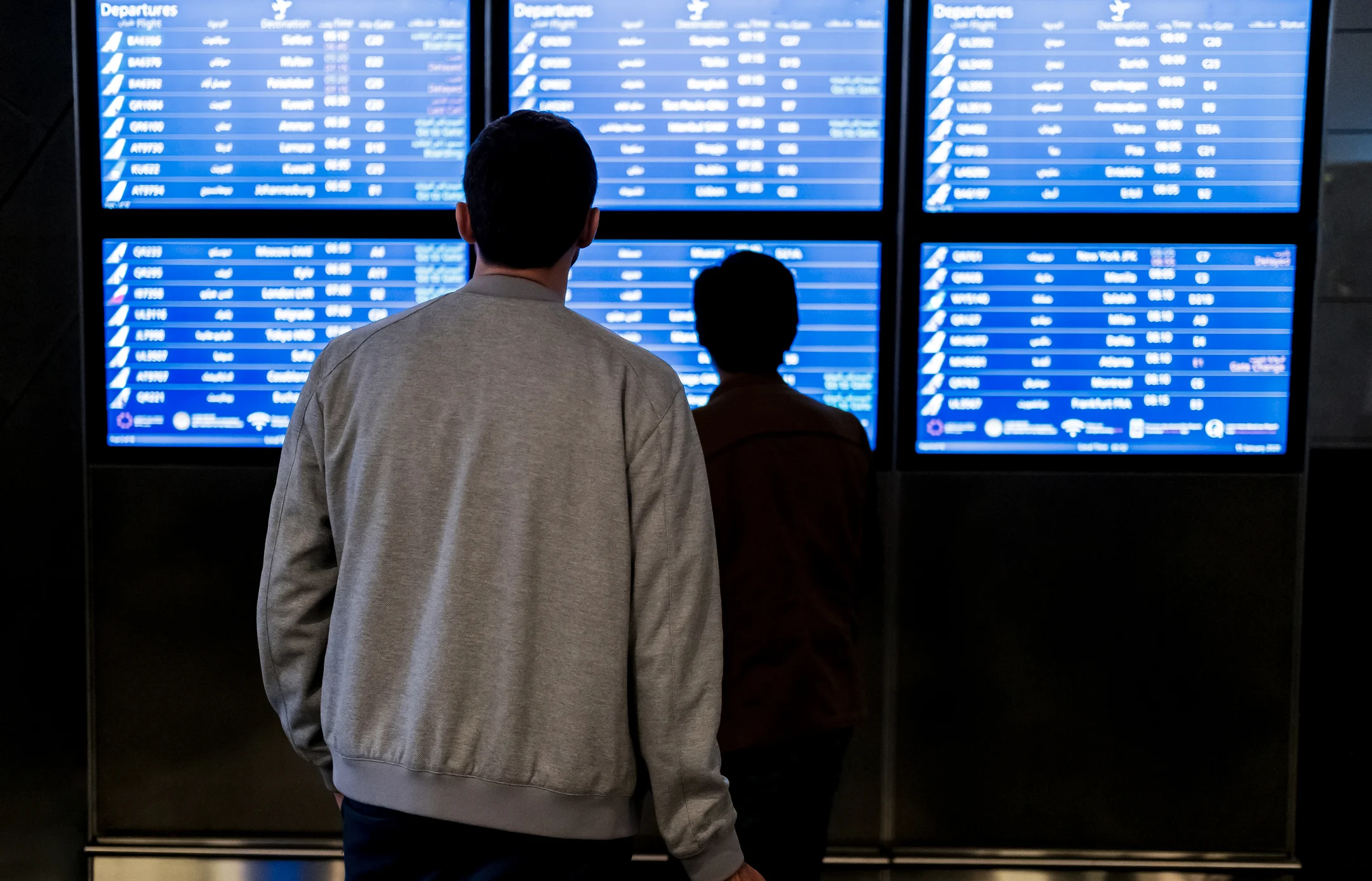 Zwei Personen betrachten die Abfluganzeigetafeln am Flughafen, auf denen Fahrgastinformationen und aktuelle Flugdaten angezeigt werden. Zwei Personen betrachten die Abfluganzeigetafeln am Flughafen, auf denen Fahrgastinformationen und aktuelle Flugdaten angezeigt werden.