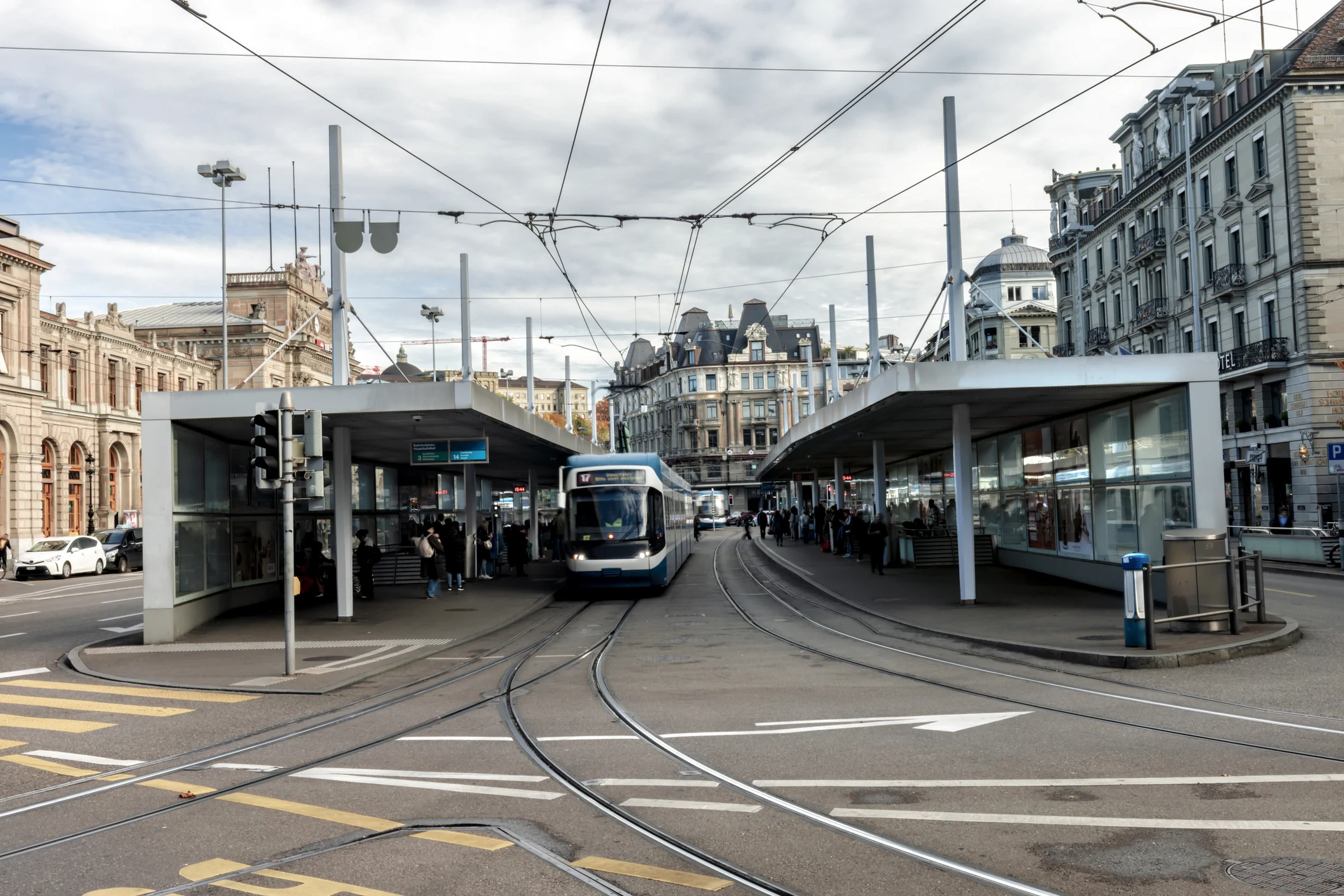 Eine blaue Straßenbahn mit Leitsystem fährt an einer Haltestelle in der Stadt ein; Fahrgäste warten vor alten Gebäuden. Eine blaue Straßenbahn mit Leitsystem fährt an einer Haltestelle in der Stadt ein; Fahrgäste warten vor alten Gebäuden.
